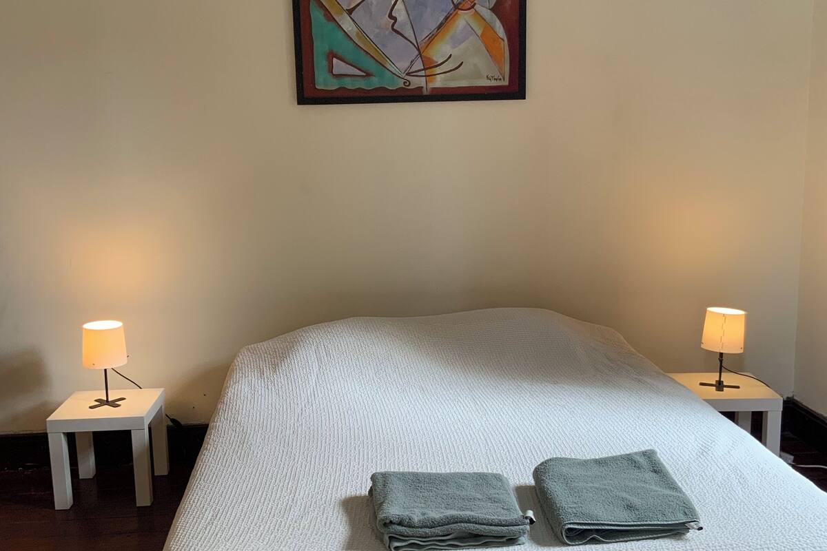 Ein ordentlich bezogenes Bett mit weißer, strukturierter Bettdecke, zwei gefaltete Handtücher und zwei Nachttische in Weiß mit je einer Nachttischlampe. Über dem Bett hängt ein farbenfrohes, abstraktes Gemälde.