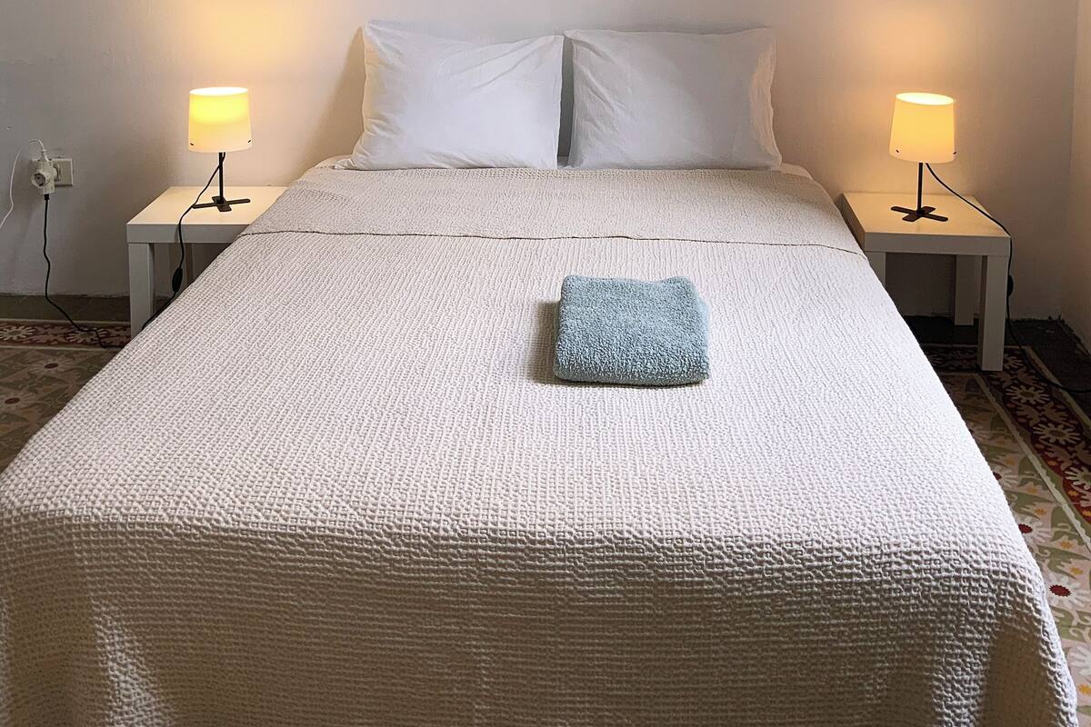 Ein ordentlich bezogenes Bett mit weißer Daunendecke und blauem Handtuch, zwei Nachttische mit Lampen und eine Steckdose an der Wand.