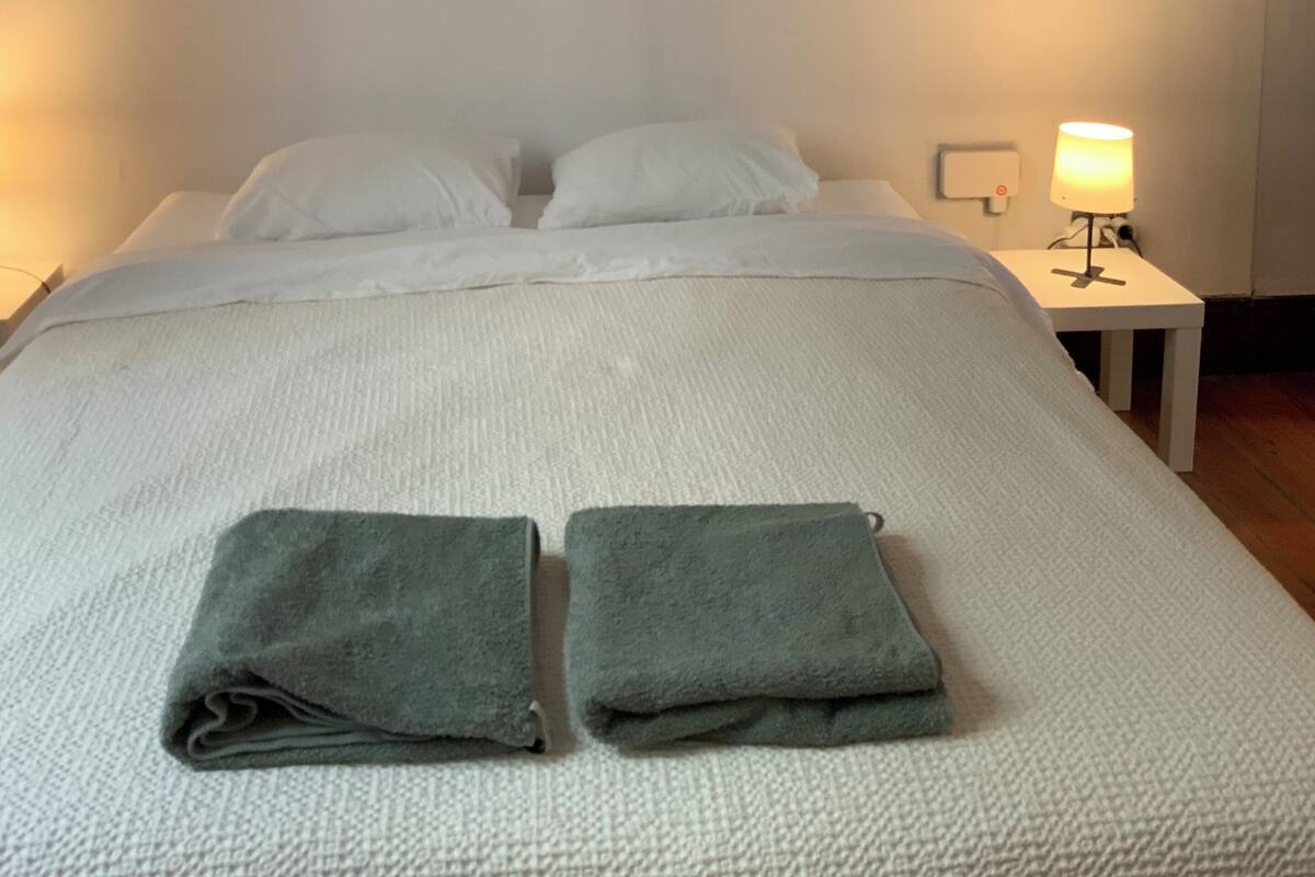 Ein ordentlich bezogenes Bett mit weißen Leinen und zwei gefalteten Handtüchern, ein Bild an der Wand, eine Nachttischlampe und ein kleiner Nachttisch.