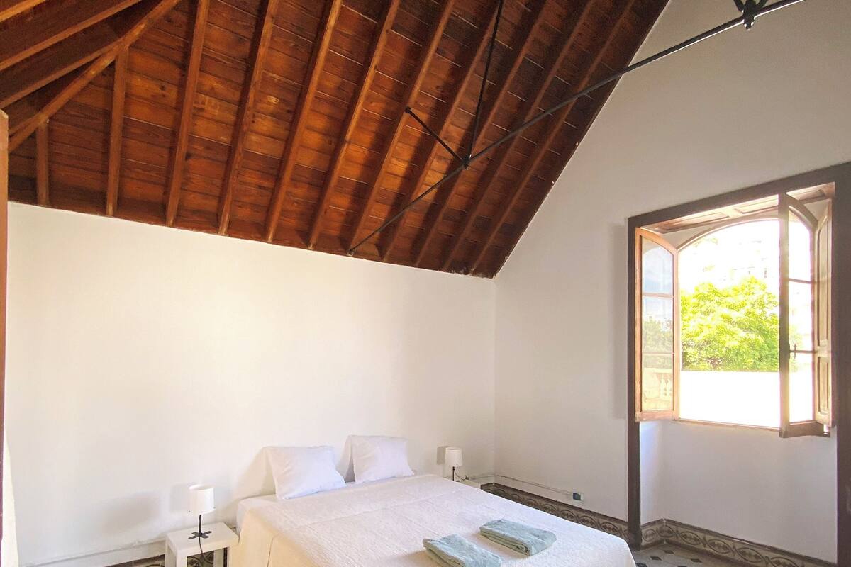Ein Schlafzimmer mit Holzbalkendecke, einem Bett mit weißer Bettwäsche, einem Nachttisch, einem Fenster mit Blick und gemusterten Bodenfliesen.