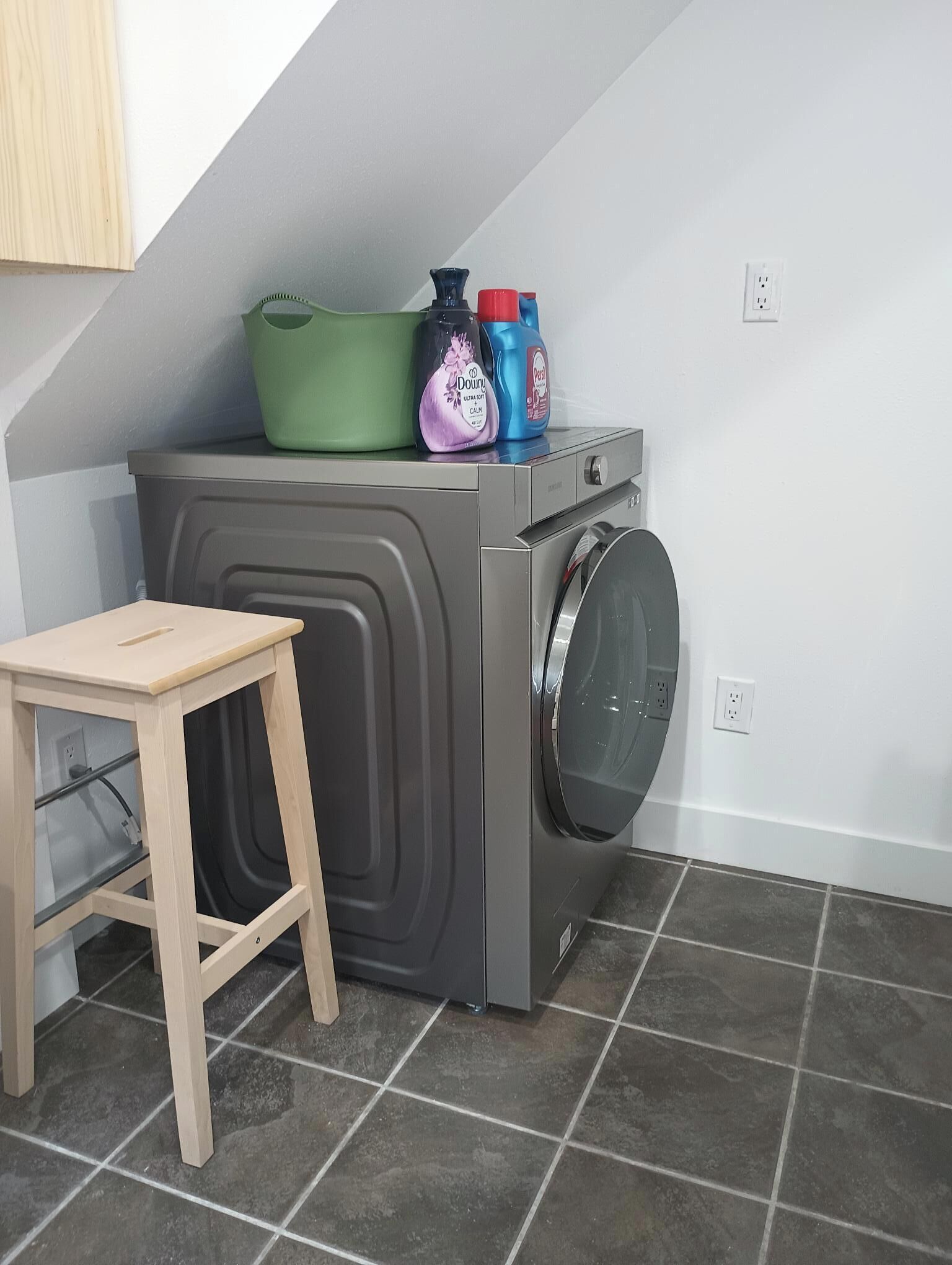 A compact laundry room with a gray washing machine, a small wooden stool, and a green basket on top.