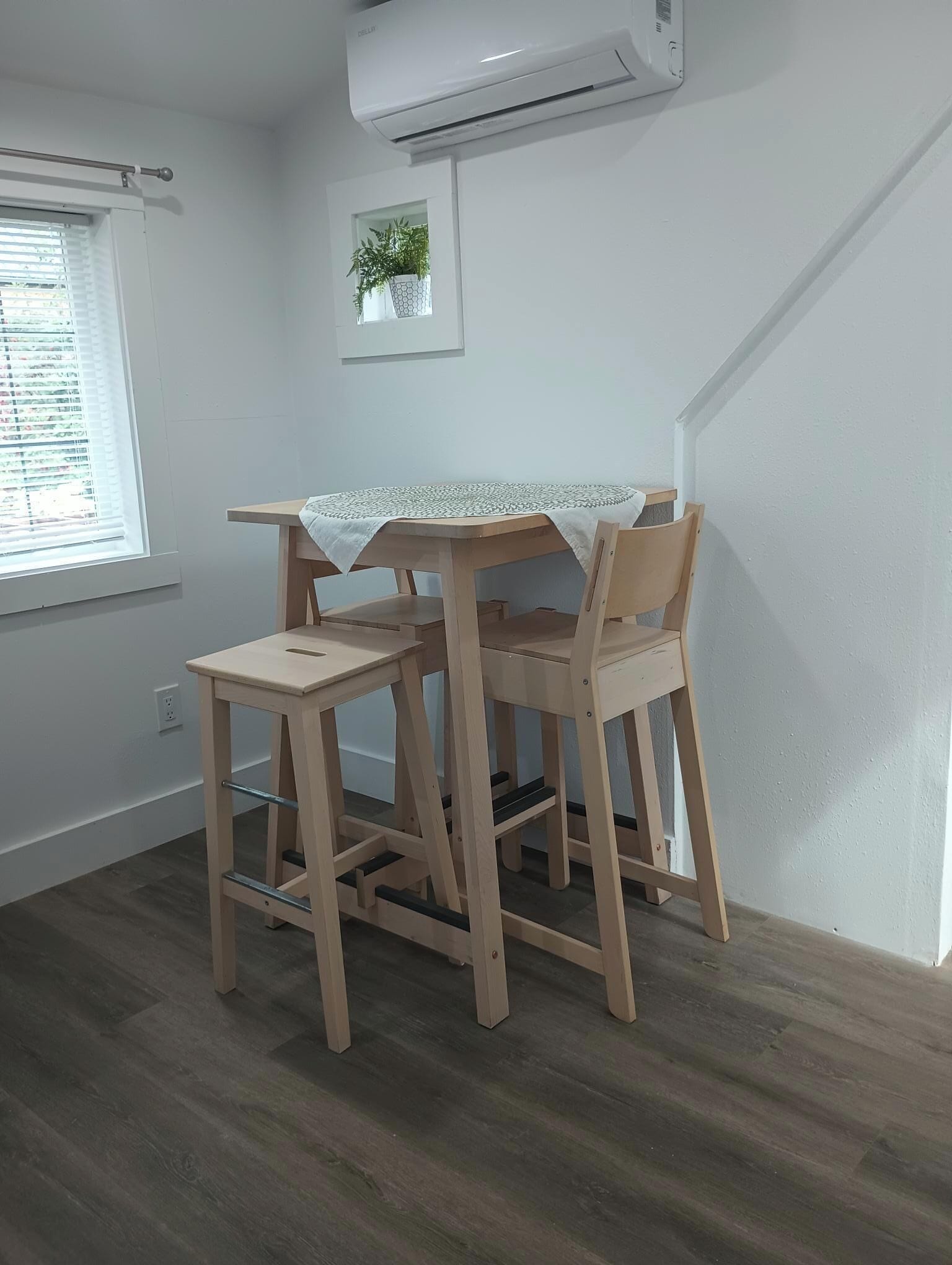 A room with a wooden table and chairs, a window with blinds, an air conditioning unit, and a small potted plant on the wall.