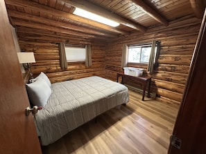 A log cabin bedroom with a bed, a nightstand, a lamp, and a window with curtains.