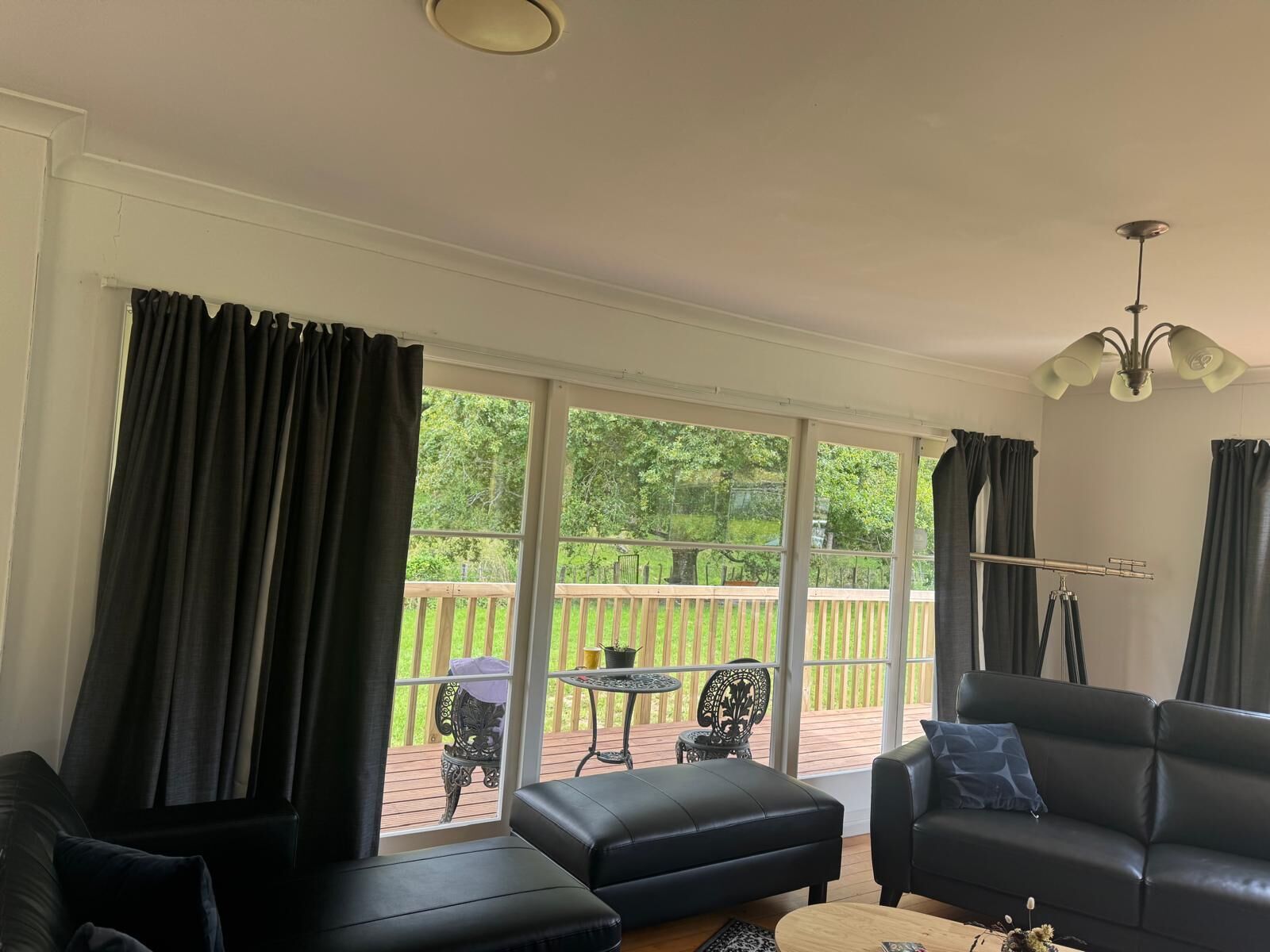 A living room with a black leather sofa, a coffee table, and a chandelier. There are large windows with curtains, and a view of a wooden deck and greenery outside.