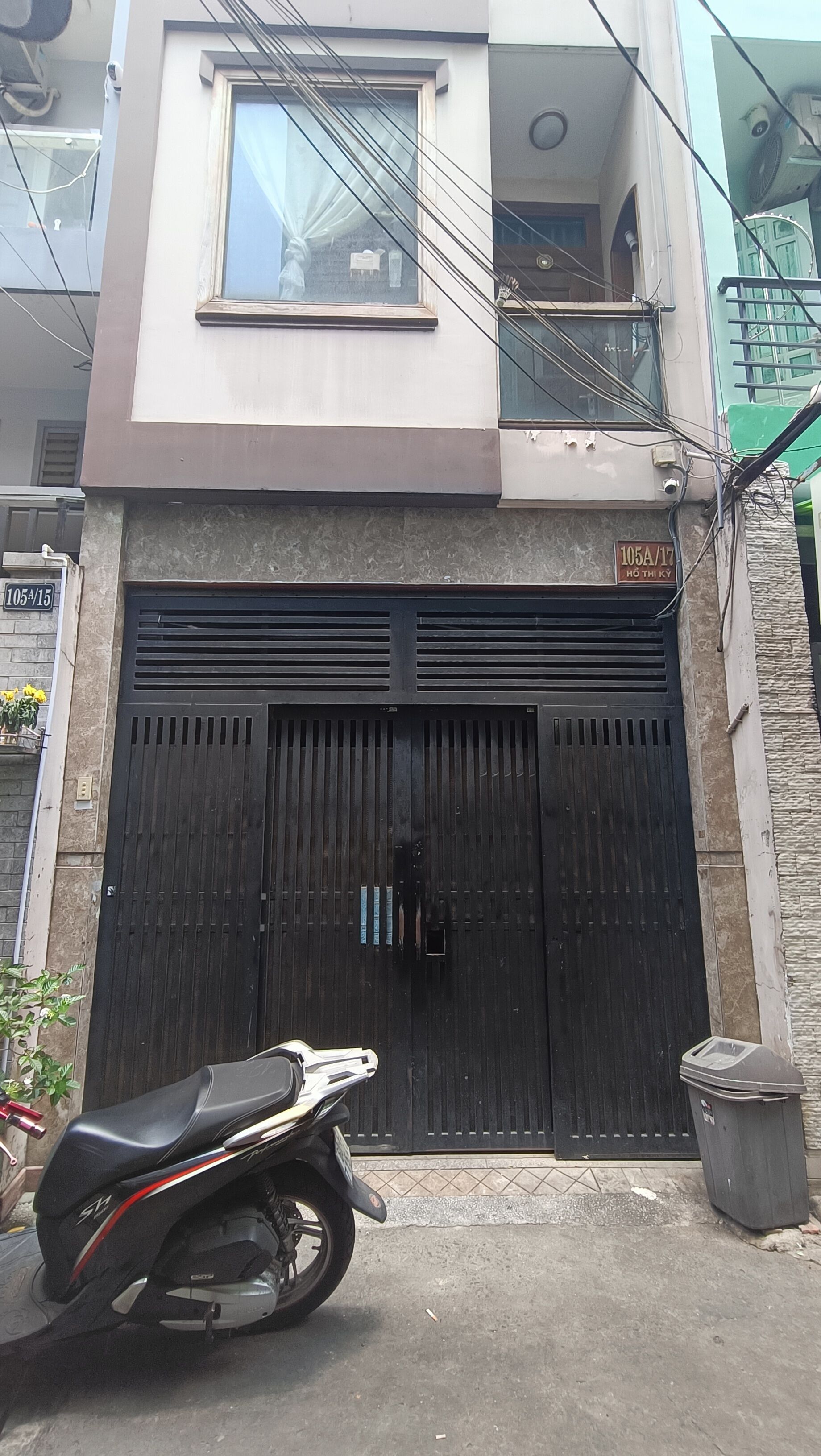 A building with a black gate, a motorcycle parked in front, and a trash bin to the right.