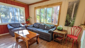 A cozy living room with a wooden table, two grey sofas, a red chair, and a view of trees through large windows. - Jackass Hostel (North Fork)