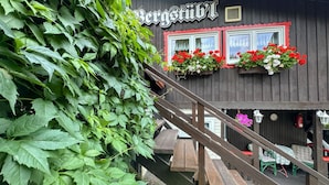 A wooden building with a sign that reads "Bergstift". There are flower boxes with red and white flowers on the windows, and a wooden staircase leading to an outdoor seating area.