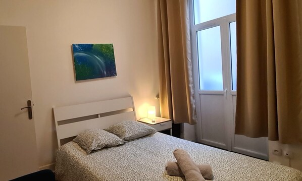 Un dormitorio con cama, almohadas, una mesita de noche, lámpara y ventana con cortinas.