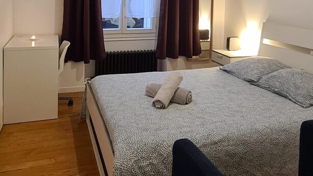 Un dormitorio con cama, mesitas de noche, una silla, una ventana con cortinas, un radiador y un cuadro en la pared.