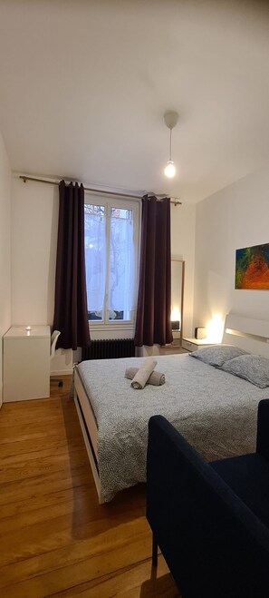 Un dormitorio con cama, mesitas de noche, una silla, una ventana con cortinas, un radiador y un cuadro en la pared.
