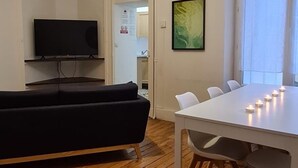 Un salón moderno con suelo de madera, un sofá negro, una mesa de comedor blanca con velas, un televisor de pantalla plana y un cuadro en la pared.