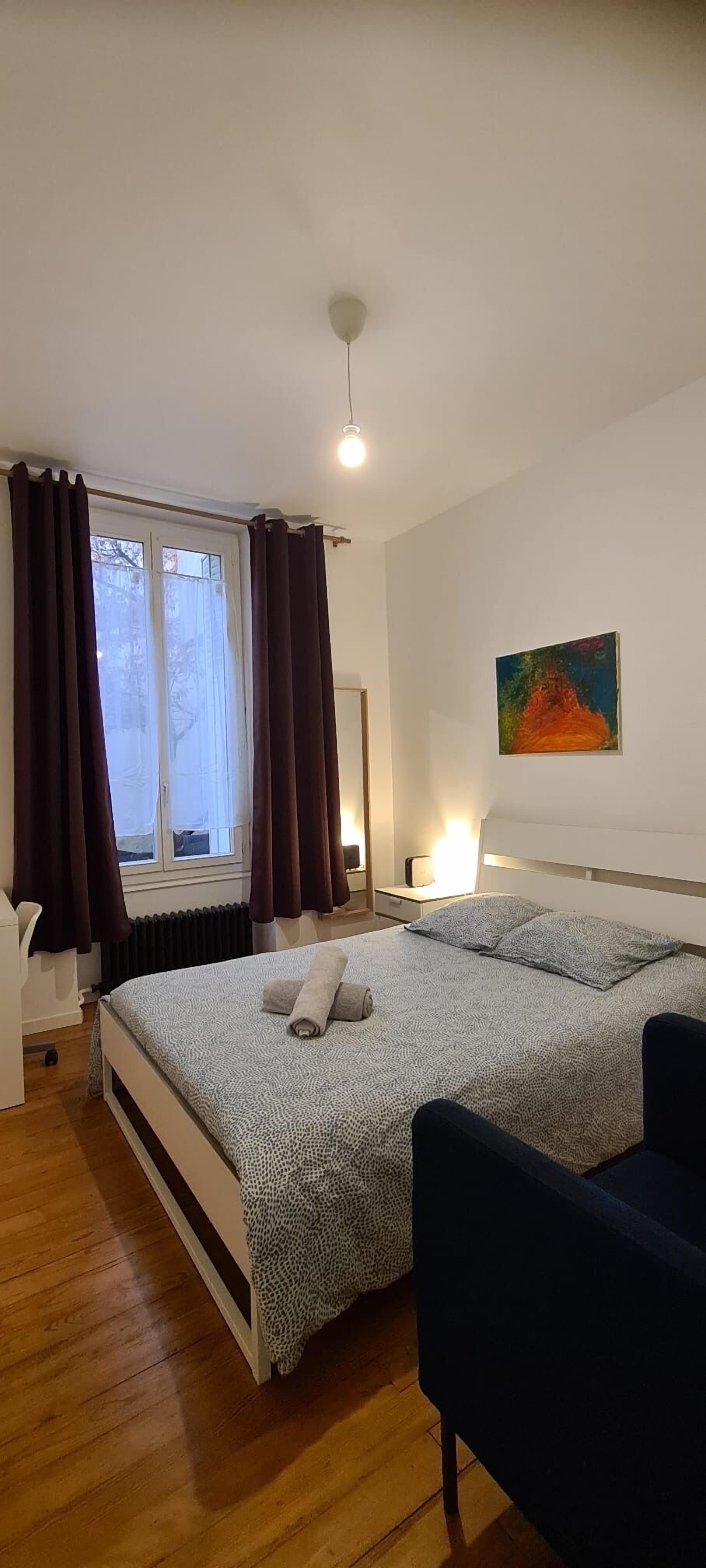 Une chambre à coucher avec un lit, une chaise, une table de chevet, un tableau et une fenêtre avec des rideaux.