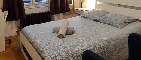 Une chambre à coucher avec un lit, une chaise, une table de chevet, un tableau et une fenêtre avec des rideaux.