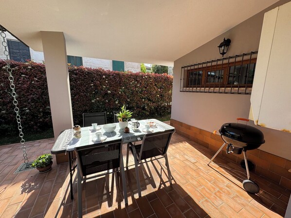 Eine Terrasse mit Esstisch, einer Hängesessel, einem Grill und einem Hecken im Hintergrund.