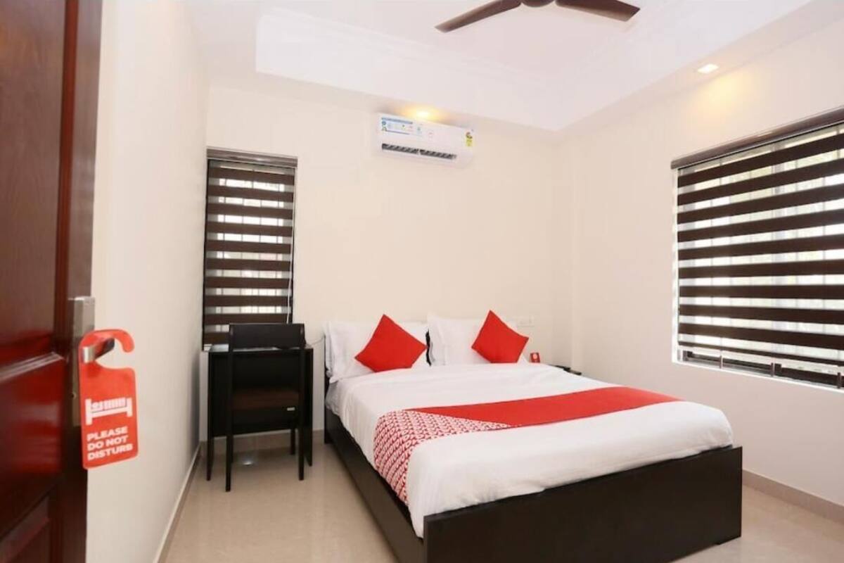 A hotel room with a bed, a desk, and a chair. There is a window with blinds, an air conditioning unit, and a door with a sign.