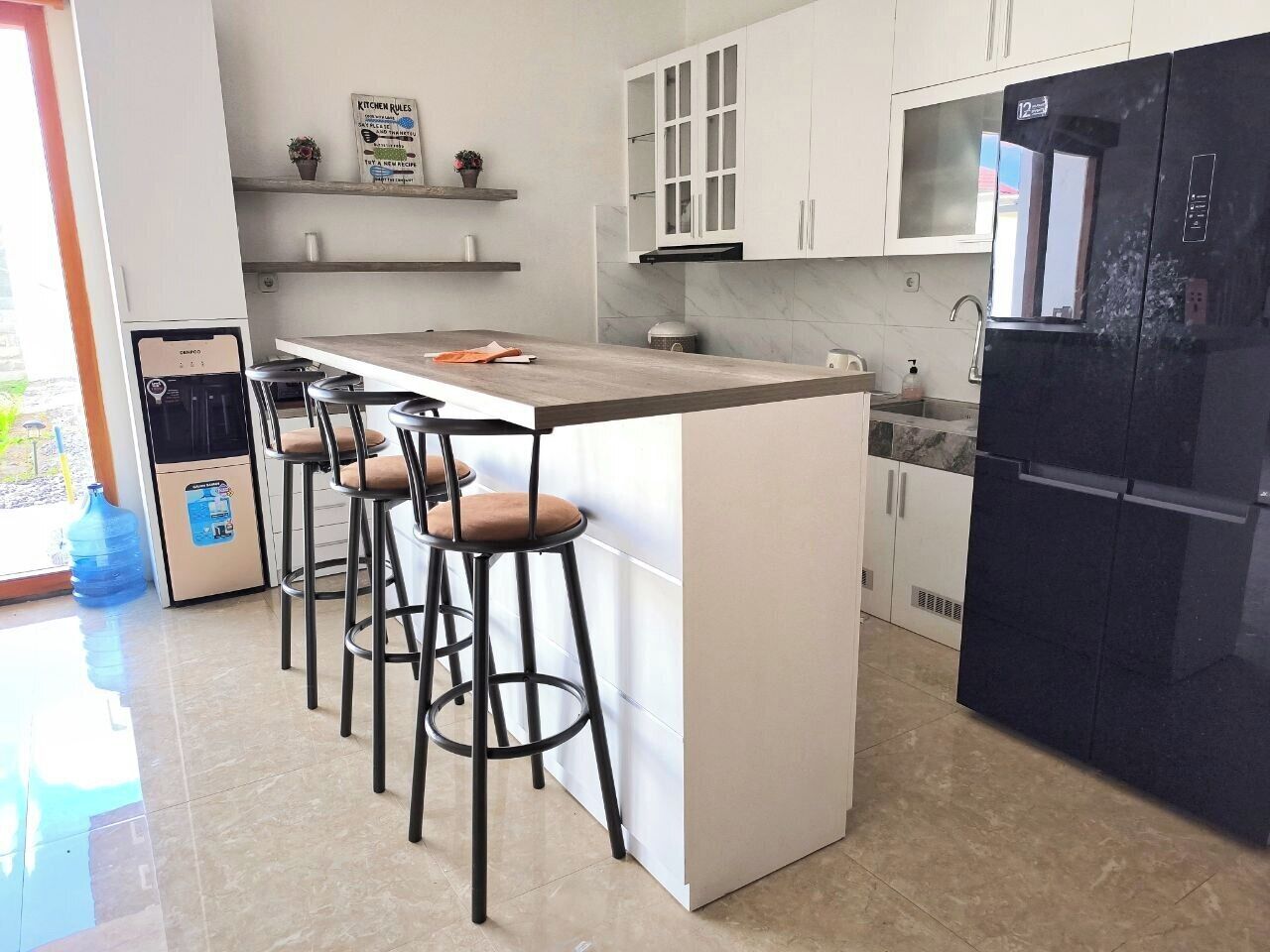 A modern kitchen with a central island, bar stools, a black refrigerator, and white cabinets.