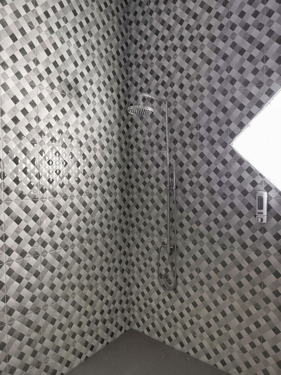 A bathroom with a diamond-patterned tile wall, a shower with a handheld showerhead, and a small window.