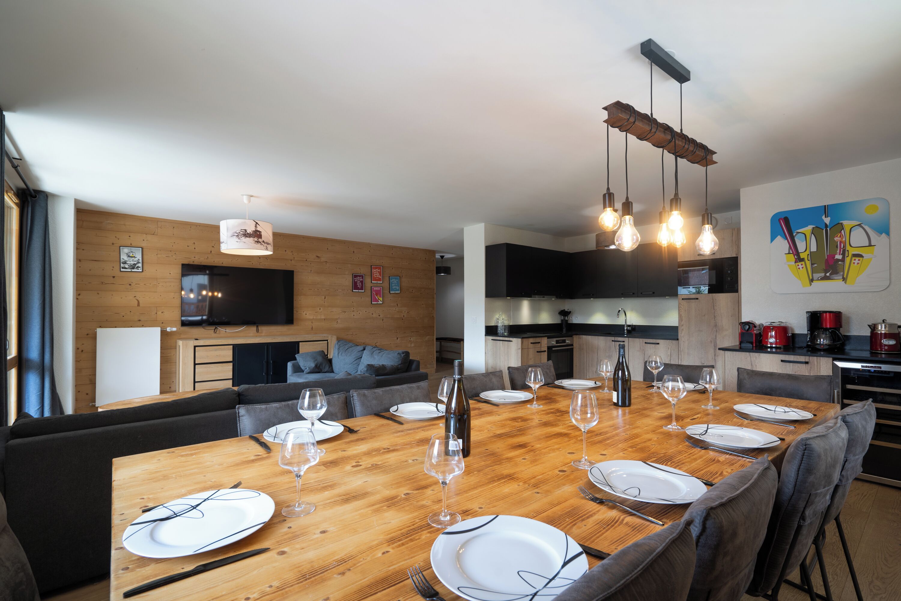 A modern dining area with a wooden table set for six, surrounded by grey upholstered chairs. A television is mounted on the wall, and a kitchen is visible in the background.