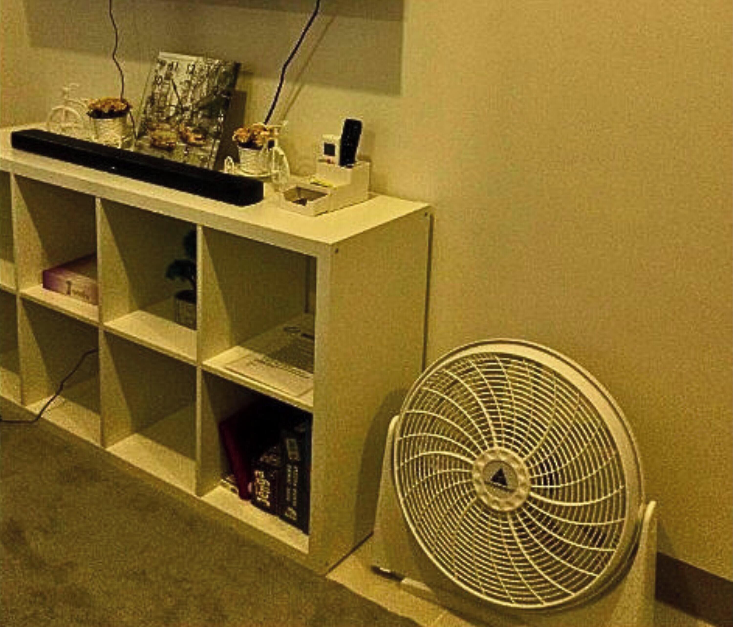 Een witte wandplank met diverse spullen, waaronder een ventilator, in een kamer met een effen muur en vloerkleed.