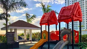 A playground with slides and climbing structures, surrounded by palm trees and a building in the background. - Rent Xpress Online US Embassy (Pasay)