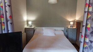 A bedroom with a bed, two bedside tables with lamps, a ceiling light, and patterned curtains. - STUDIO FOR RENT IN PRATS DE MOLLO, Pyrénées-Orientales.  THE MOST BEAUTIFUL VILLAGE IN FRANCE.  (Prats-de-Mollo-la-Preste)
