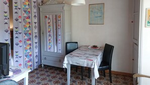 A dining area with a table, chairs, a cabinet, patterned curtains, a hanging light, and a framed picture on the wall. - STUDIO FOR RENT IN PRATS DE MOLLO, Pyrénées-Orientales.  THE MOST BEAUTIFUL VILLAGE IN FRANCE.  (Prats-de-Mollo-la-Preste)