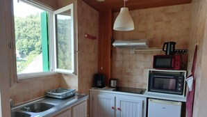 A compact kitchen with a window, a white refrigerator, a microwave, and a kettle on the counter. - STUDIO FOR RENT IN PRATS DE MOLLO, Pyrénées-Orientales.  THE MOST BEAUTIFUL VILLAGE IN FRANCE.  (Prats-de-Mollo-la-Preste)