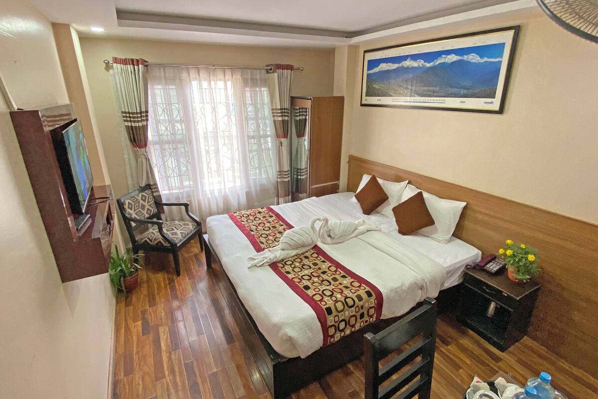 A hotel room with a neatly made bed, a desk with a telephone and a plant, a chair, and a picture of the Himalayas on the wall.
