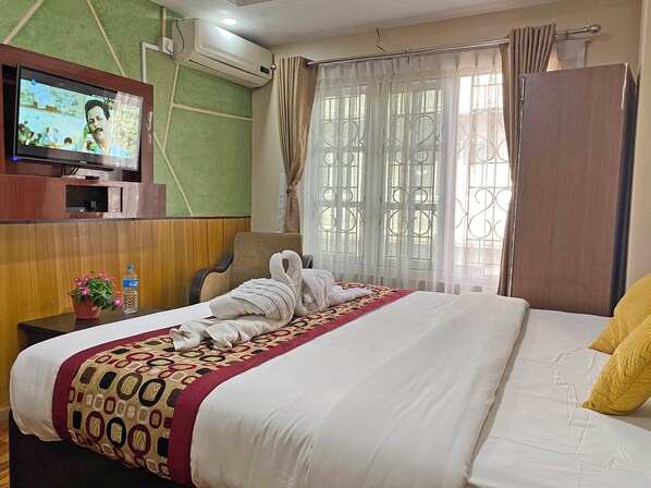 Une chambre d’hôtel avec un lit, un téléviseur fixé au mur, un appareil de climatisation, une fenêtre avec des rideaux et une petite table sur laquelle se trouvent une plante et une bouteille.
