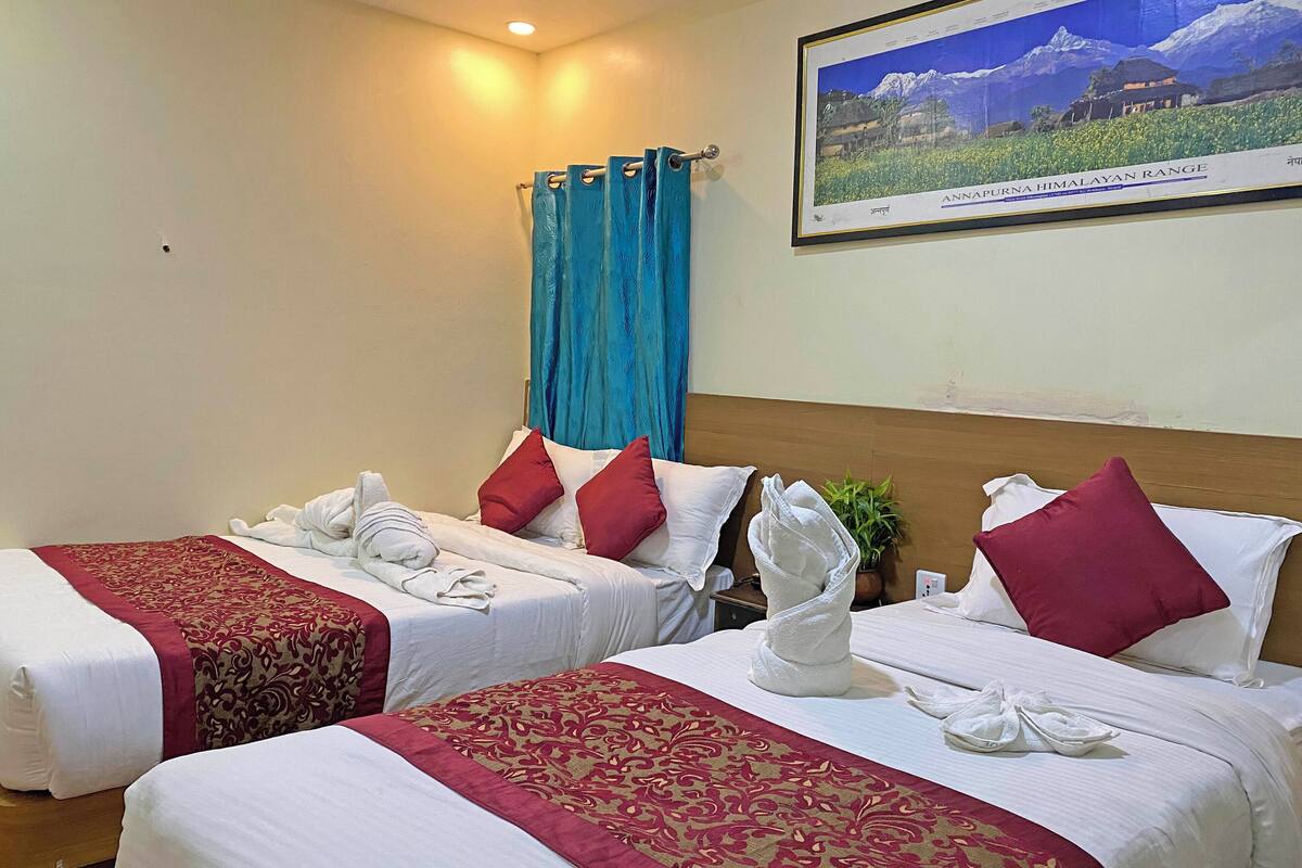 A hotel room with two beds, each with red and white bedding, a blue curtain, and a framed picture of the Annapurna Himalayan Range on the wall.