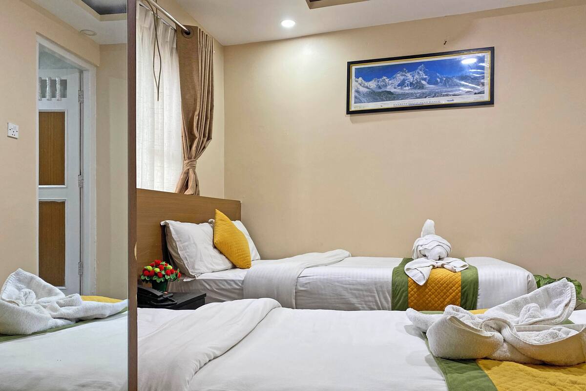 A hotel room with two single beds, a wooden headboard, a framed picture of a mountain, and a small table with a vase of flowers.