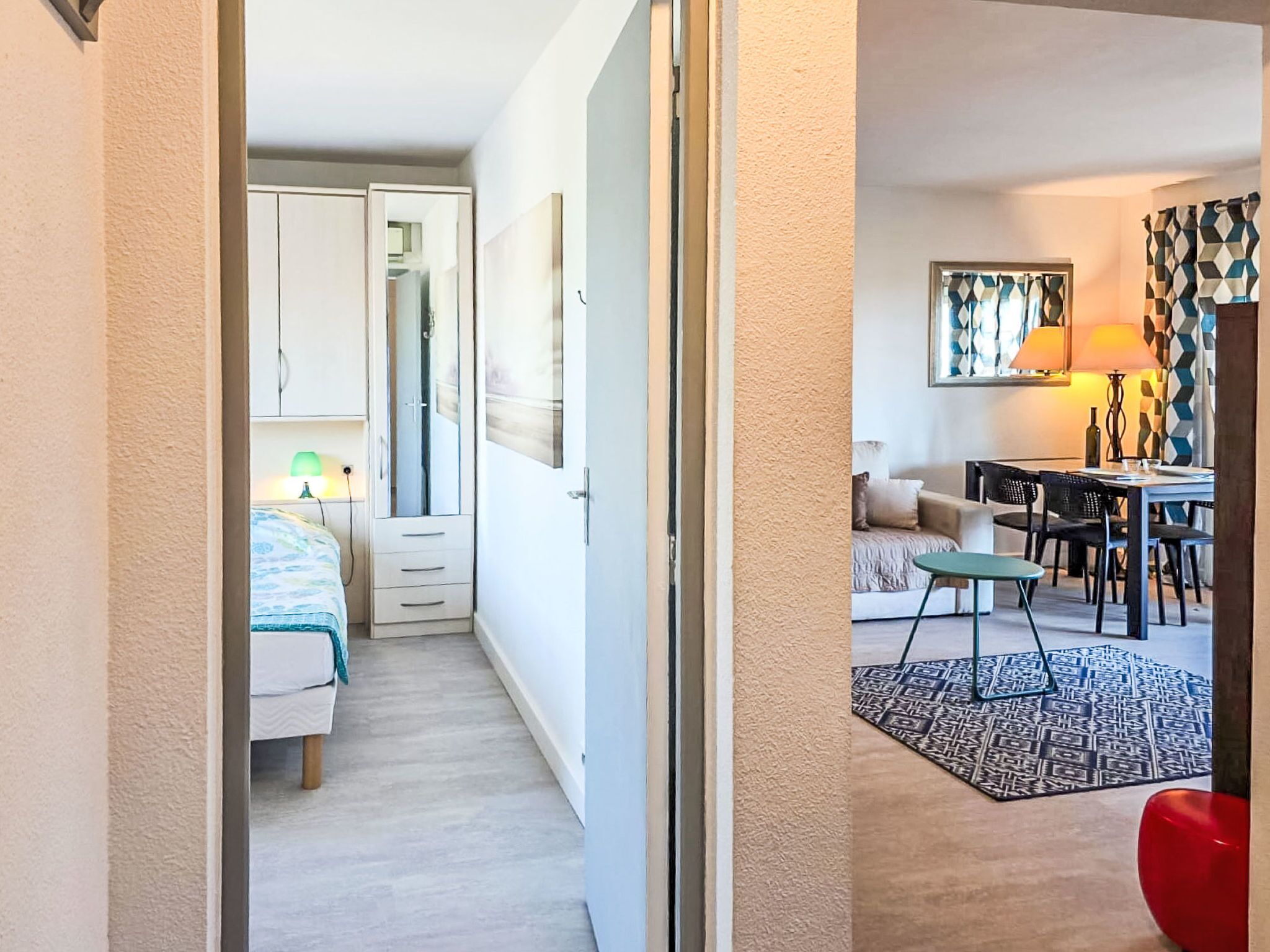 Ett hotellrum med en säng, ett skrivbord, en stol, en röd stol, en mönstrad matta och en tavla på väggen.
