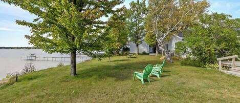 A grassy area with a bench and a tree, a dock extending into a body of water, and a house in the background.