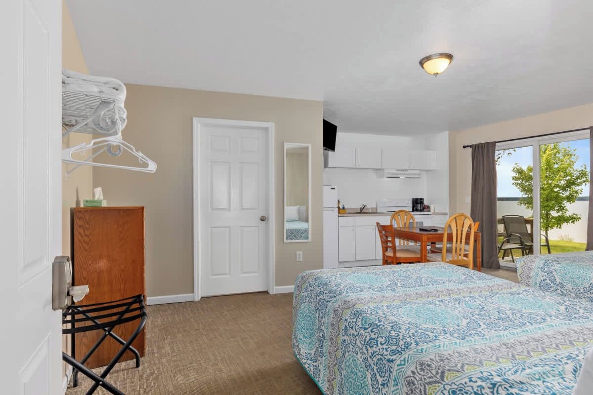 A hotel room with a bed, a desk, a chair, a kitchenette, and a view of the outdoors.