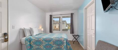 A bedroom with a bed, a desk, a chair, a TV mounted on the wall, and a window with curtains.