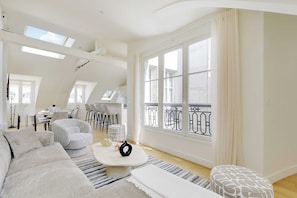 A spacious living area with a large window, a sofa, a coffee table, and a dining table with chairs. - Apt a/c 3br/6p - Saint-lazare/les Champs Elysee (Paris)