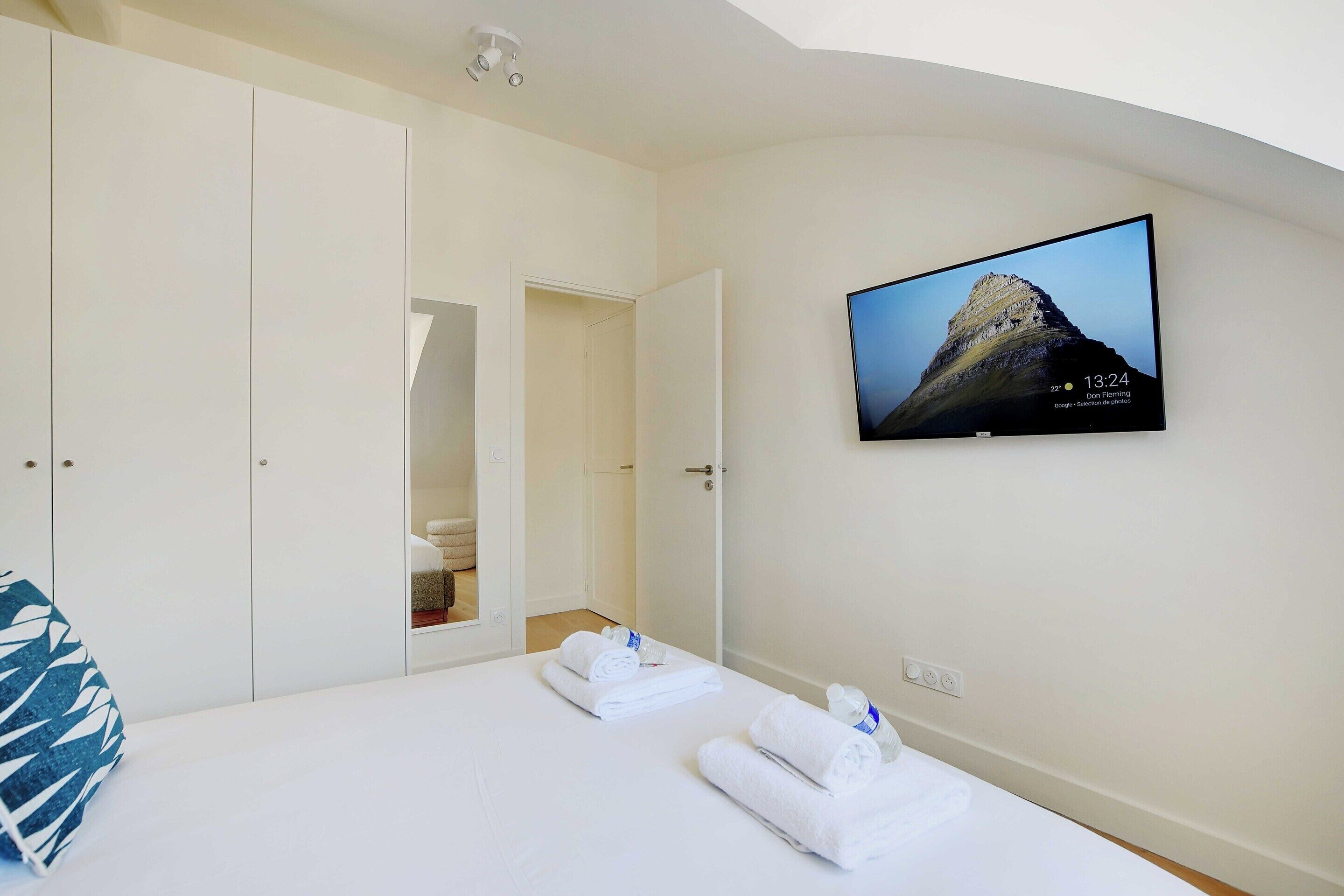 A hotel room with a bed, a TV mounted on the wall, a wardrobe, and a bathroom visible through an open door.