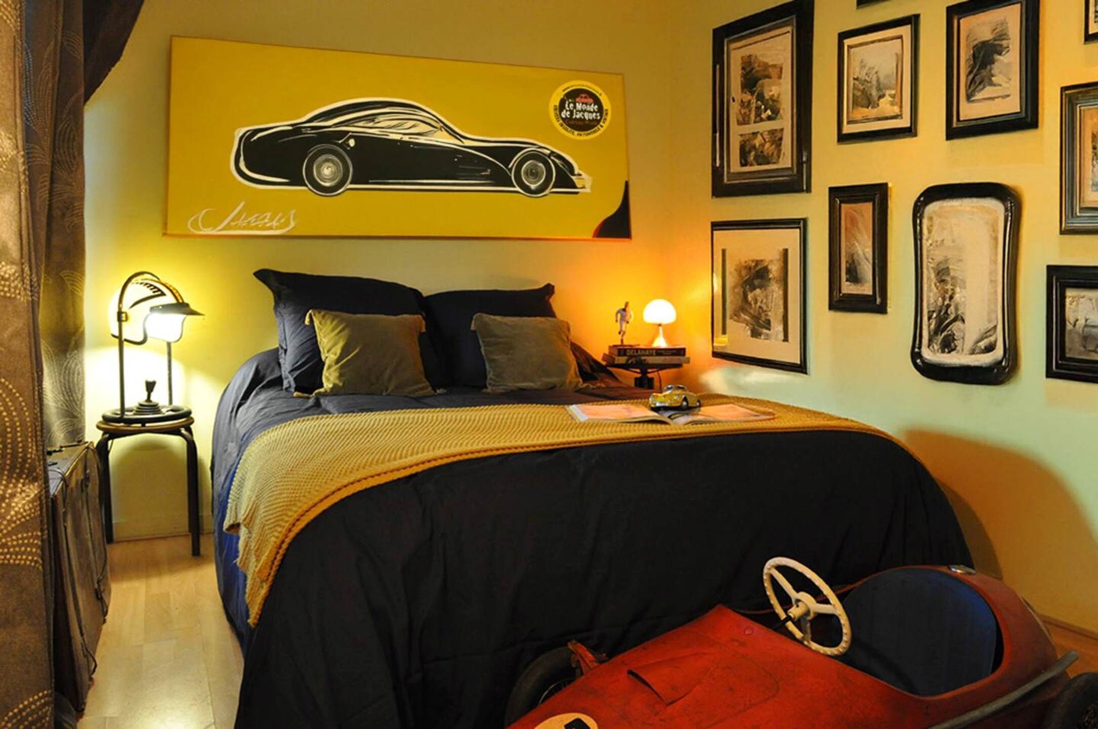 Un dormitorio con cama, almohadas, una mesita de noche con lámpara, un cuadro en la pared y un coche de juguete sobre la cama.