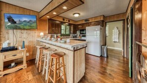 A kitchen with wooden cabinets, a central island with stools, a refrigerator, and a painting of a bird in flight.