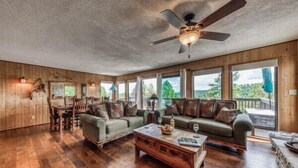 A spacious living room with wooden floors, a ceiling fan, and large windows offering a view of the outdoors.