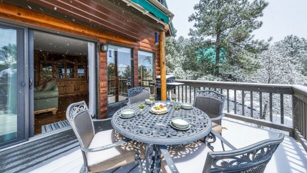 A wooden deck with a round table and chairs, a glass door leading inside, and snow-covered trees in the background.