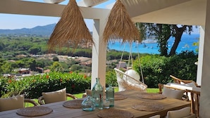 A wooden table with woven placemats, wicker chairs, and a hanging chair with a white cushion, overlooking a scenic landscape with greenery and a view of the sea.