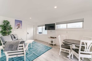 A modern living room with a sofa, a dining table with chairs, a TV mounted on the wall, and a large window with blinds.