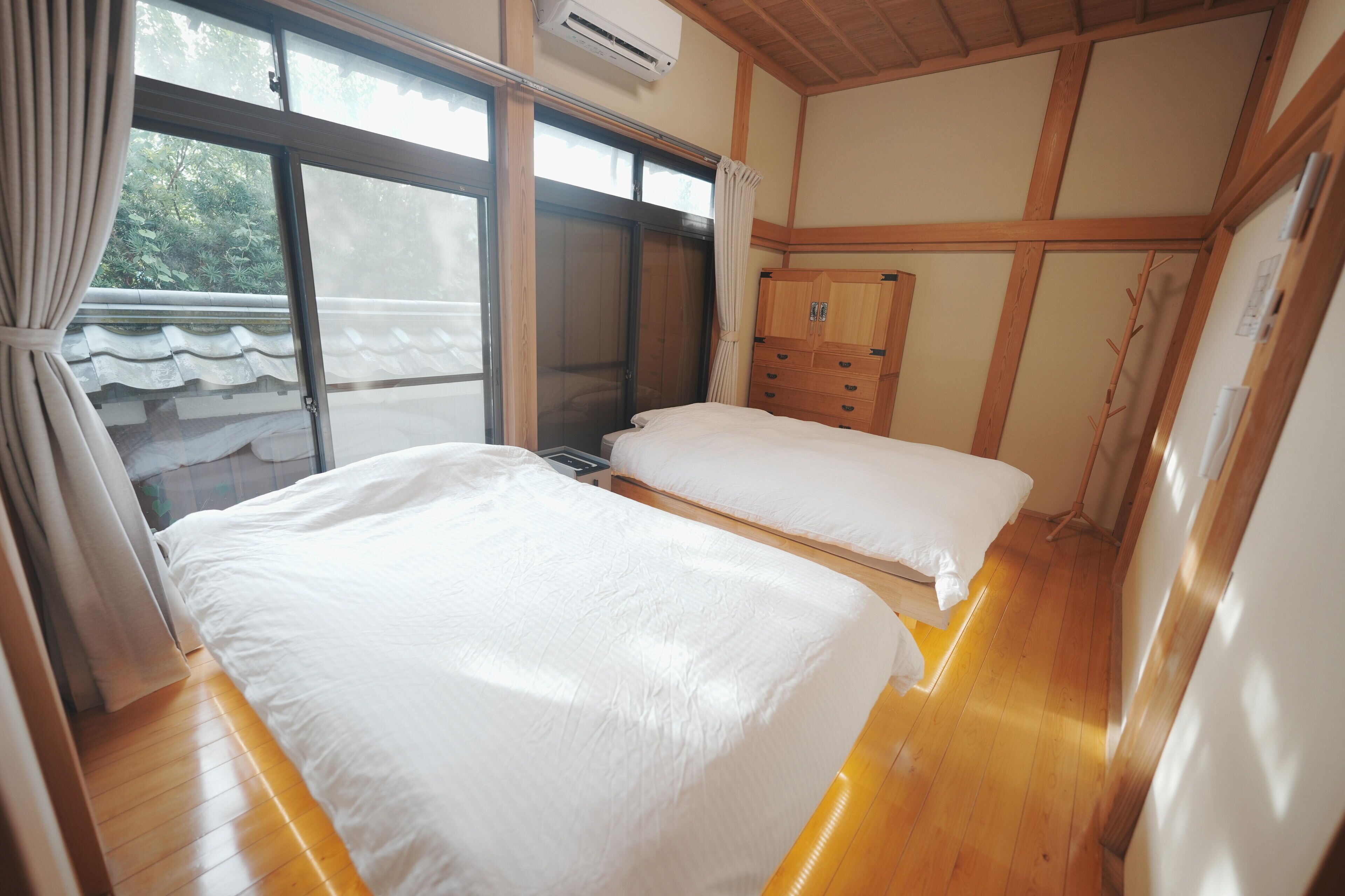 A room with two beds, wooden flooring, a wardrobe, and a sliding door leading to a balcony.