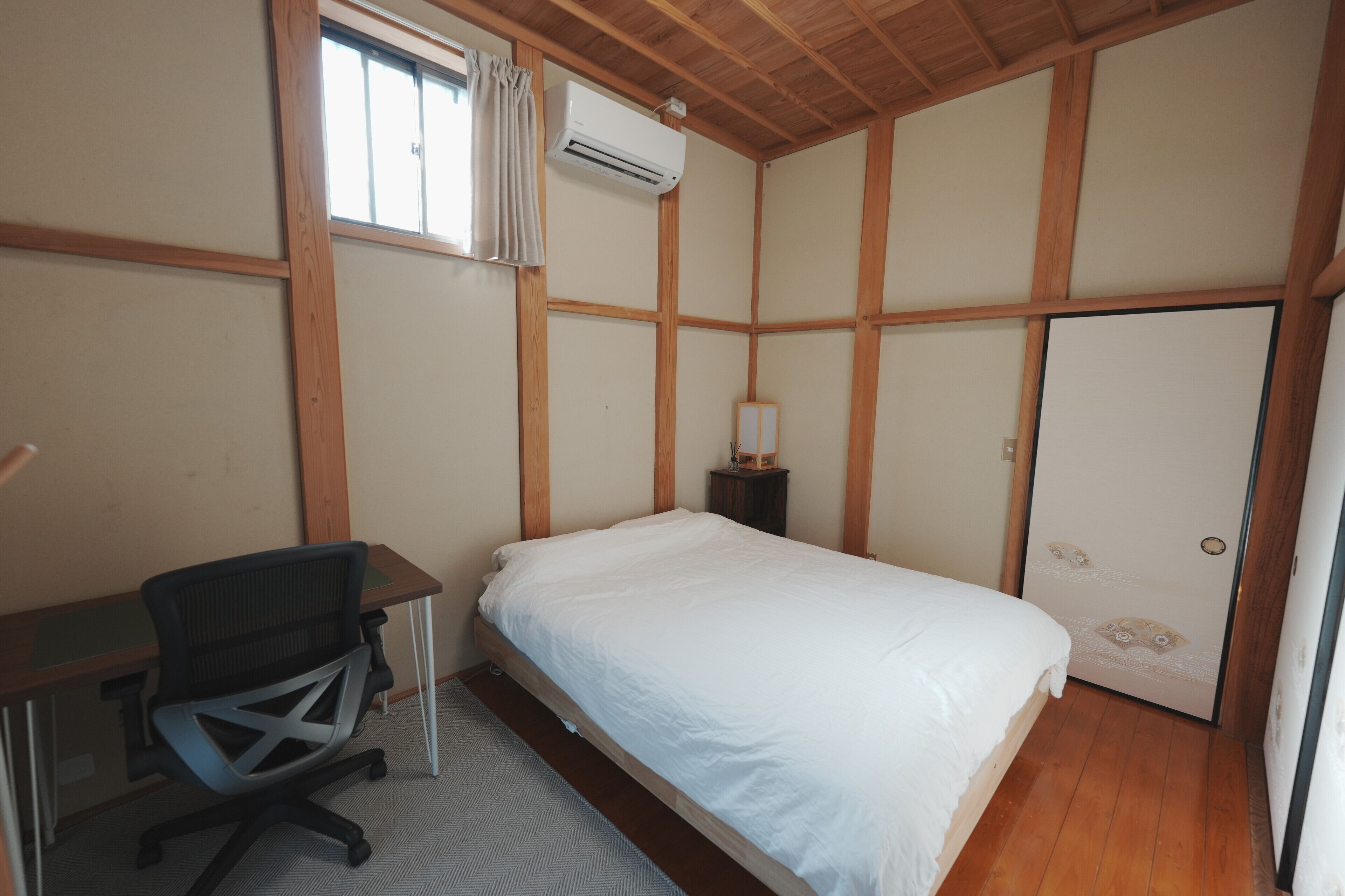 A bedroom with a bed, desk, chair, and an air conditioning unit.
