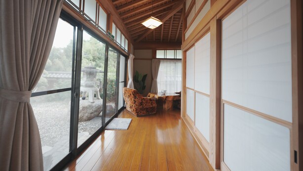 Una habitación con suelo de madera, ventanas amplias con cortinas, una silla tradicional y vistas a un jardín.