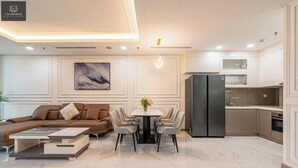 A modern living room with a sofa, coffee table, and dining area with chairs. There is a kitchen with a refrigerator and cabinets.