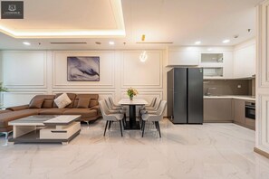 A modern living room with a sofa, coffee table, and dining area with chairs. There is a kitchen with a refrigerator and cabinets.