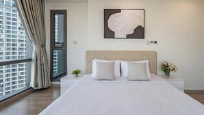 A hotel room with a large bed, a headboard, a window with curtains, a wall-mounted artwork, and a nightstand with a vase of flowers.