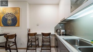 A modern kitchen with a white countertop, stainless steel sink, and a coffee maker. There are two wooden chairs and a small table with a glass.